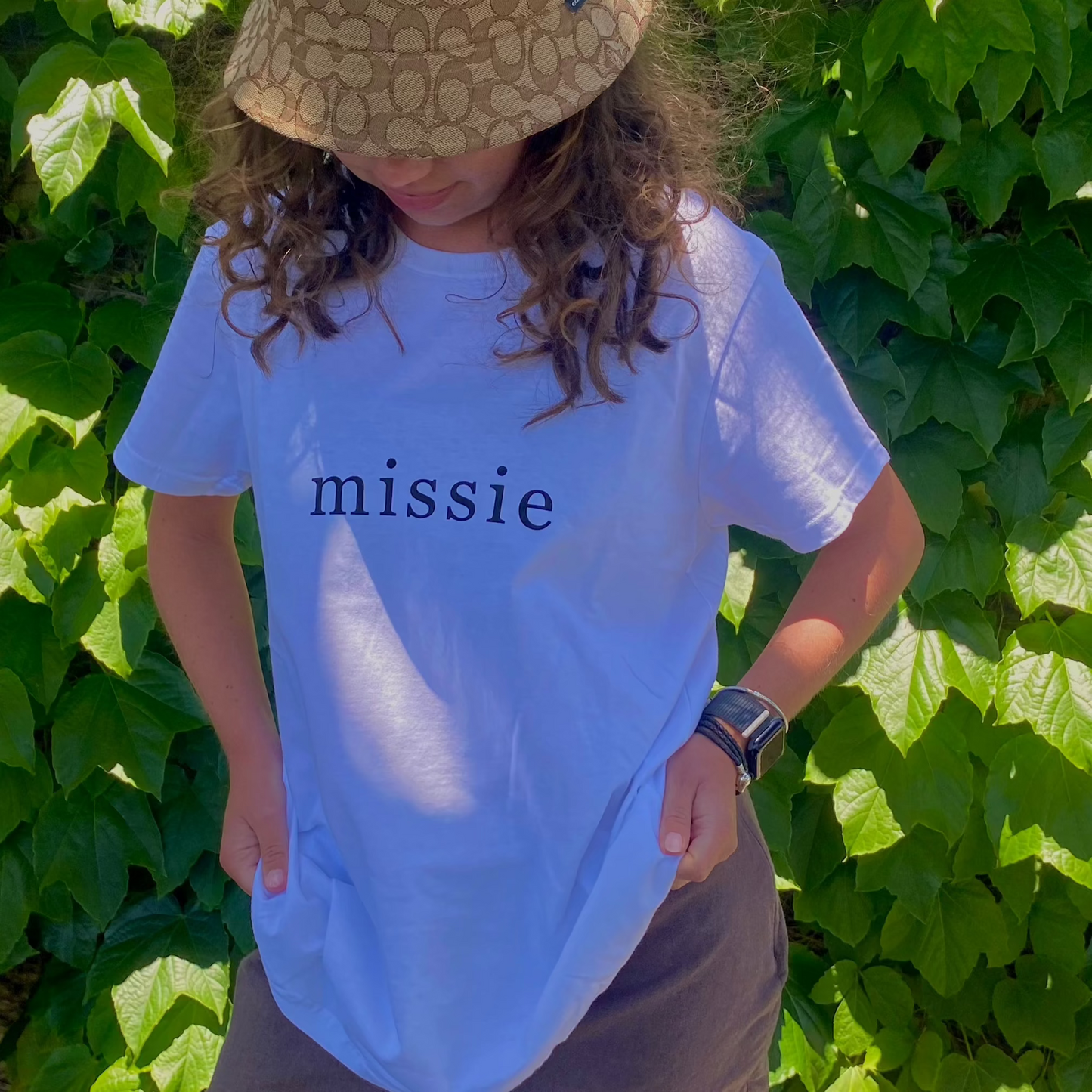 Person wearing a white t-shirt with 'missie' text in front of green foliage