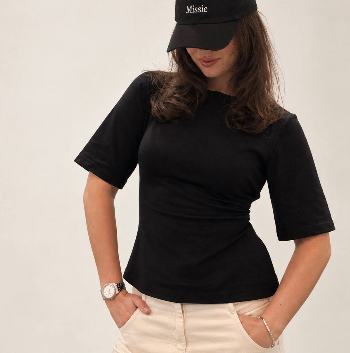 A woman wearing a fitted black short-sleeve t-shirt, cream-colored pants, and a black baseball cap with the word 'Missie' embroidered in white on the front, posed against a light gray studio background.