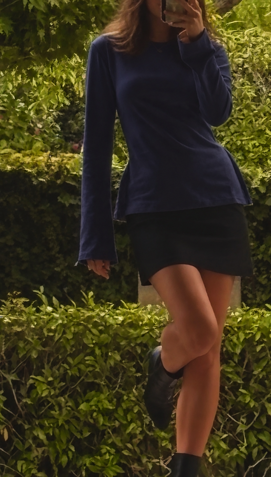A person wearing a navy blue long-sleeve top and a black mini skirt, standing outdoors in front of green hedges and taking a mirror-style photo with a phone.
