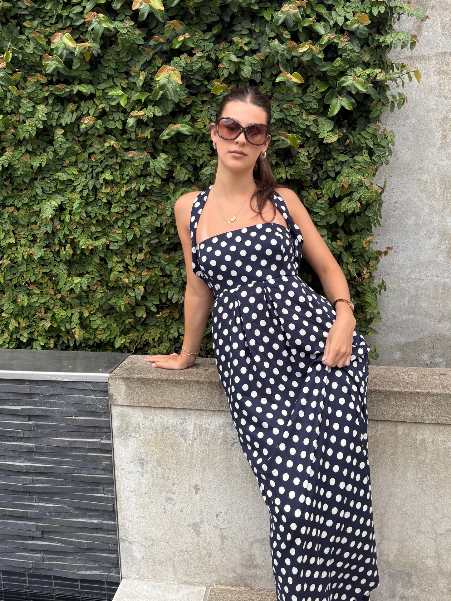 Woman in a polka dot maxi dress standing against a green hedge.