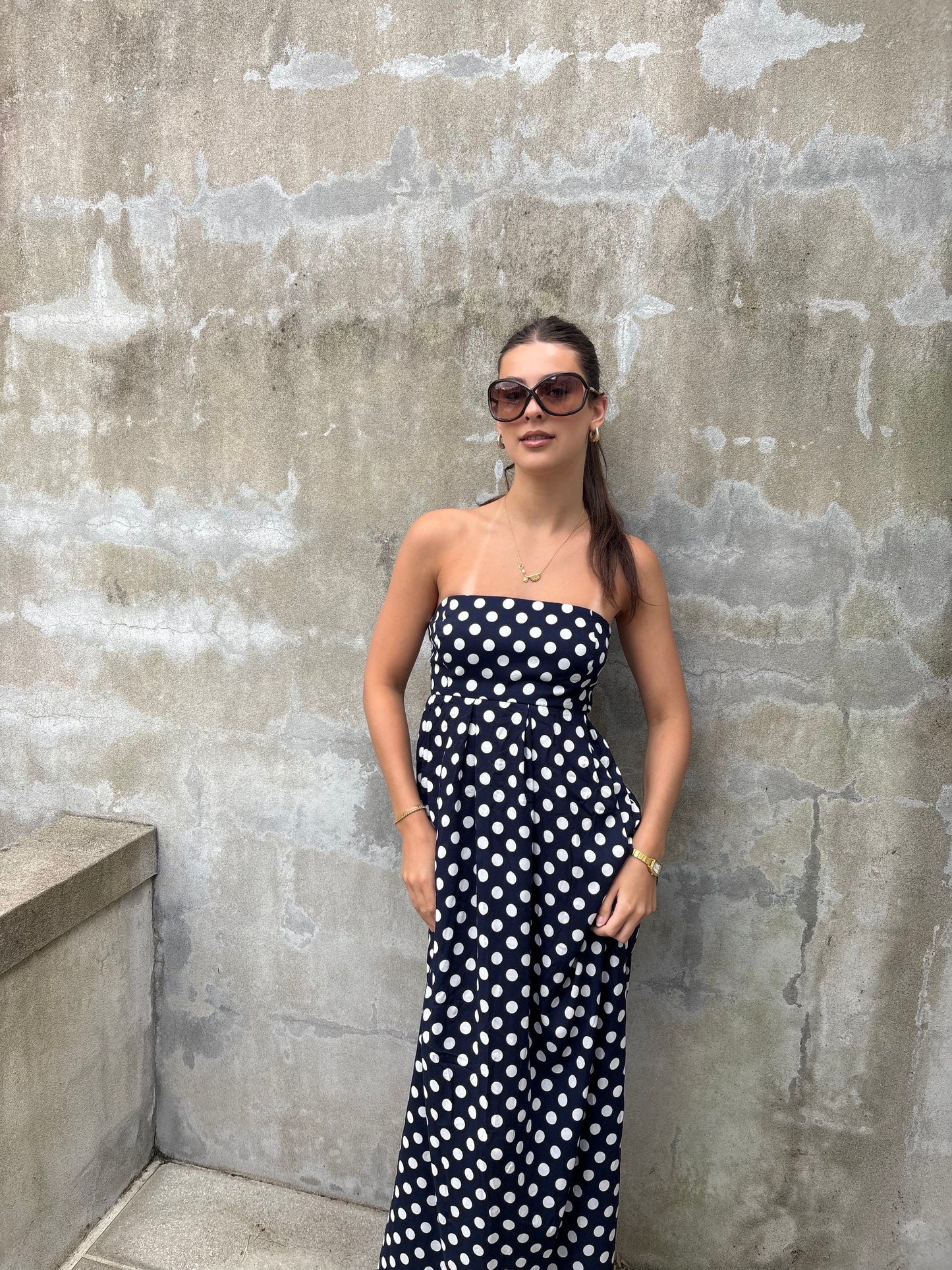 Woman in a strapless navy & white polka dot maxi dress standing against a textured wall.