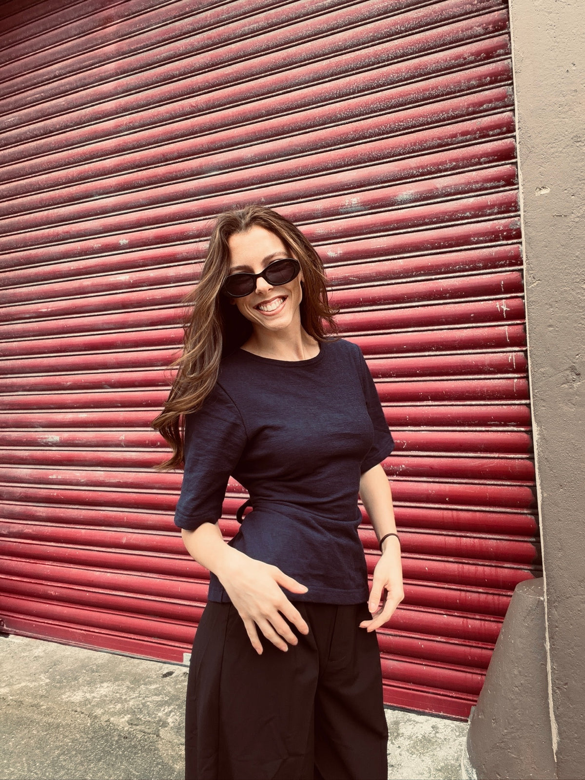 A woman wearing a dark navy blue short-sleeve top and black pants stands in front of a red metal shutter door.