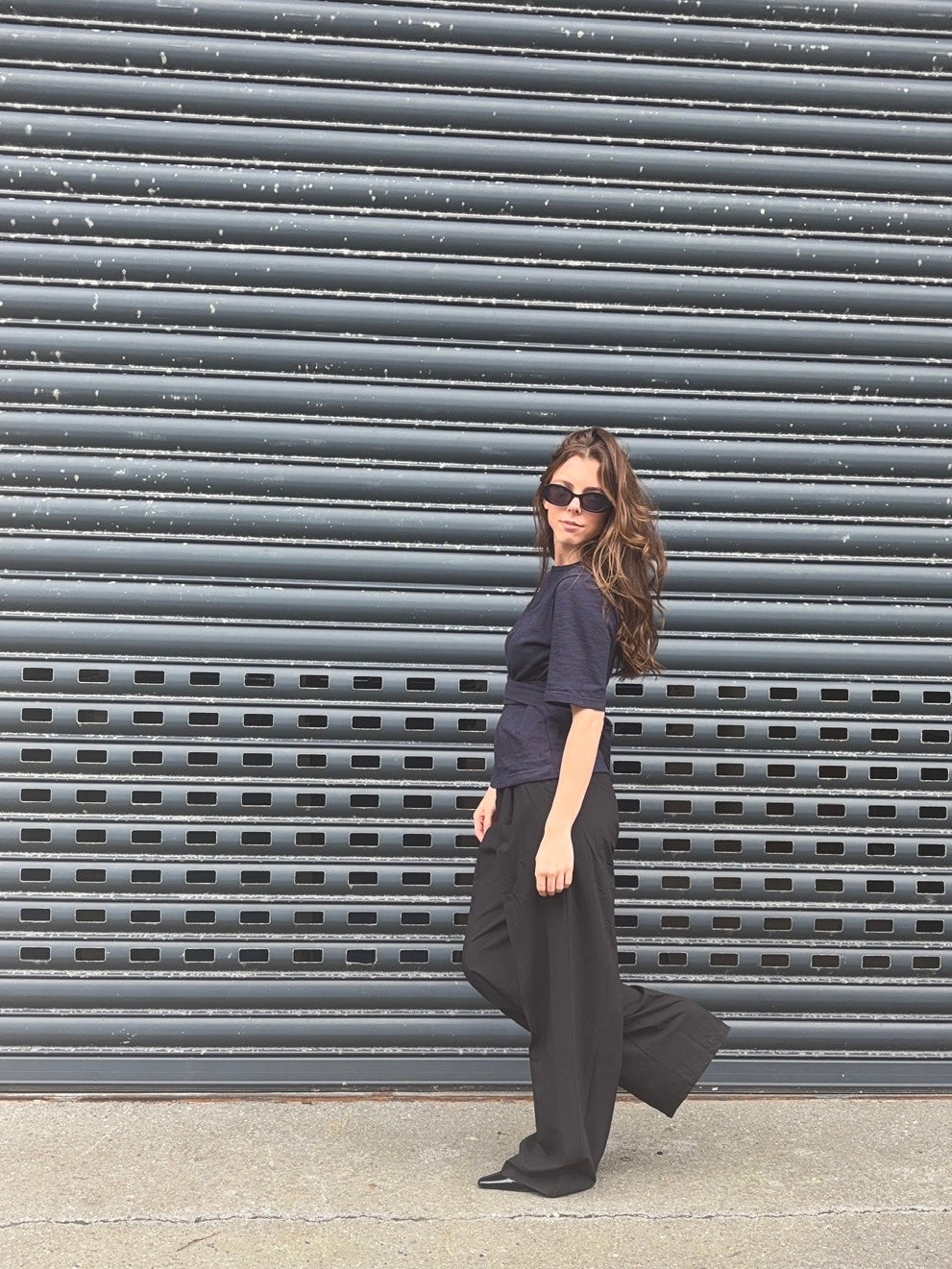 A woman wearing a dark navy blue ruffled short-sleeve top and black wide-leg pants walks in front of a gray metal roll-up door.