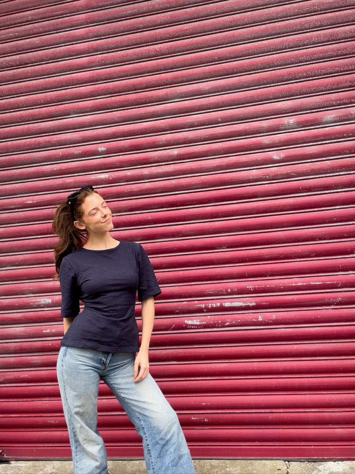 A woman wearing a dark navy blue short-sleeve t-shirt and light blue jeans stands in front of a large, textured red metal roll-up door.