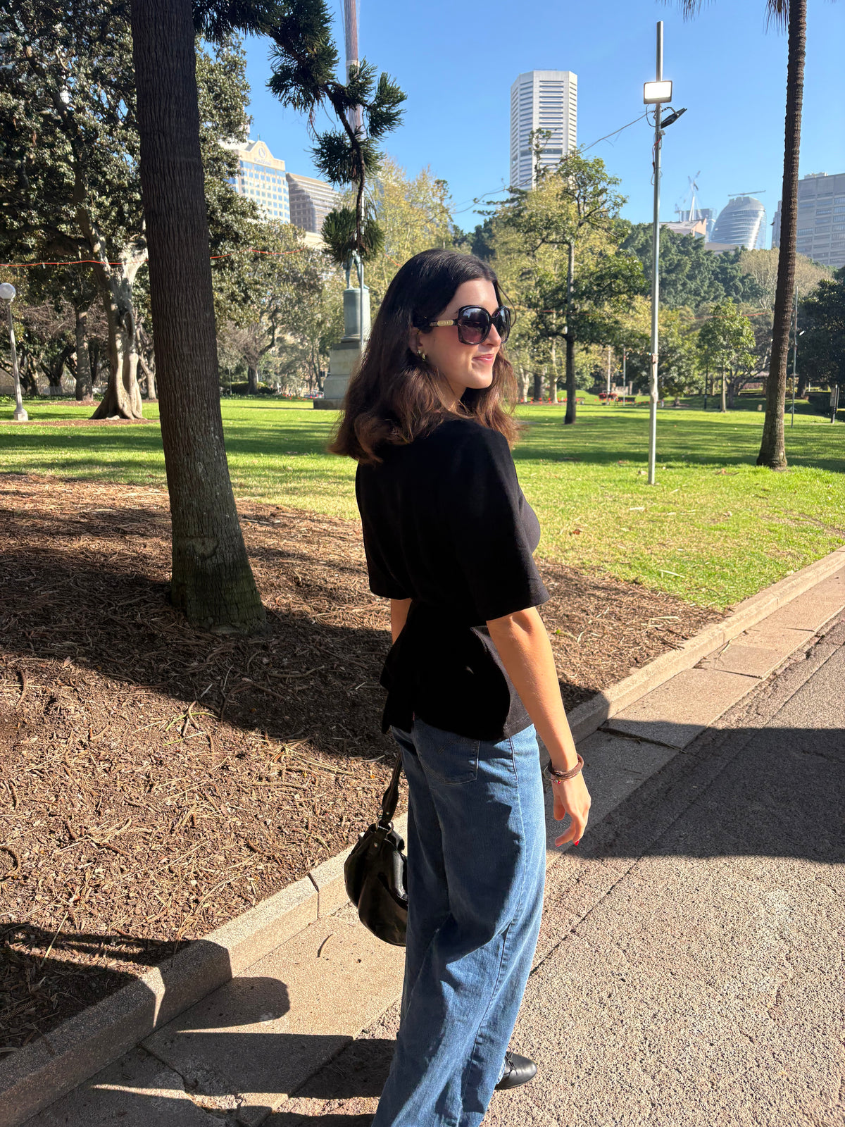 A woman wearing a black short-sleeve t-shirt and blue jeans stands on a paved path in a sunny park with trees, grass, and city buildings in the background.