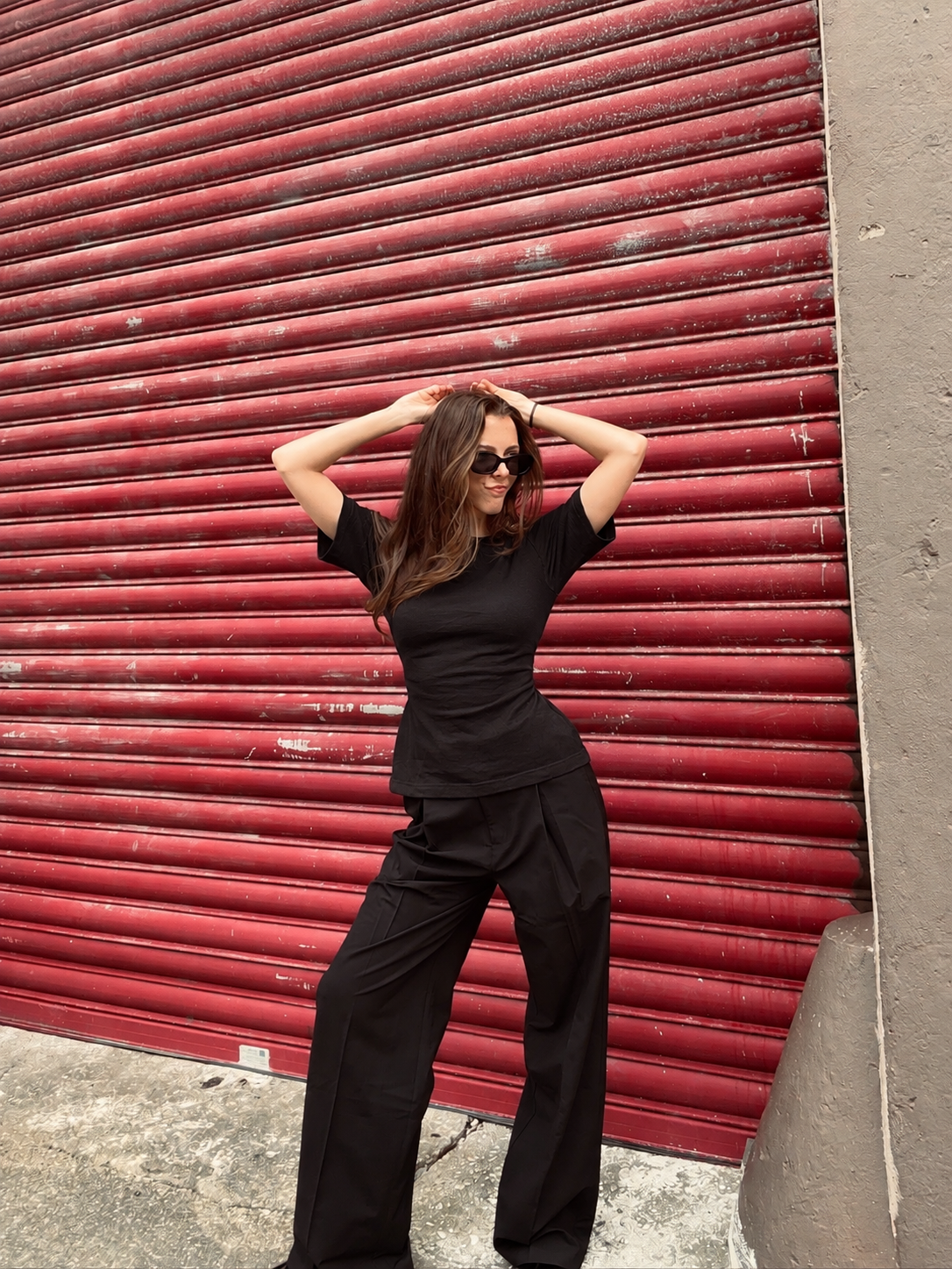 A woman posing in front of a red corrugated metal shutter, wearing a black short-sleeve top.