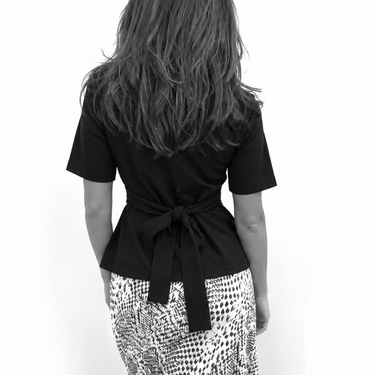 A woman shown from behind wearing a black short-sleeve wrap-style top with a tied waist and a black-and-white snakeskin print skirt, against a plain white background.