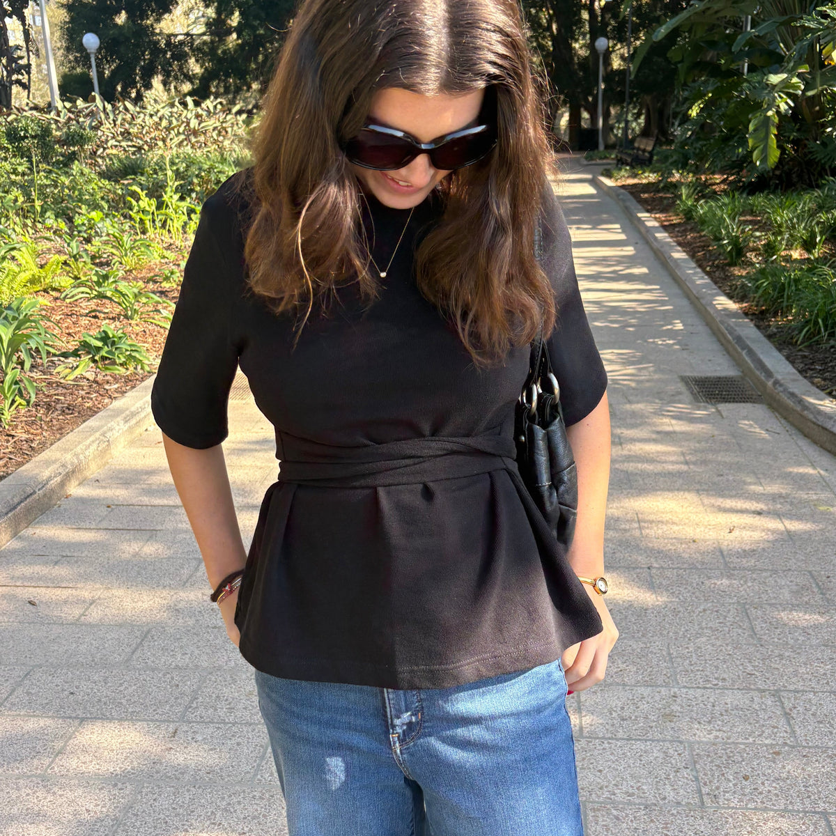 A woman wearing a black short-sleeve belted top and blue jeans walking on a paved path in a park.